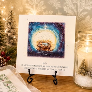 May include: A square white card on a black metal stand featuring a watercolor illustration of a baby in a manger under a bright star against a dark blue starry sky. Below the image, the text reads: Day 25. For God so loved the world that he gave his one and only Son, that whoever believes in him shall not perish but have eternal life. - John 3:16.