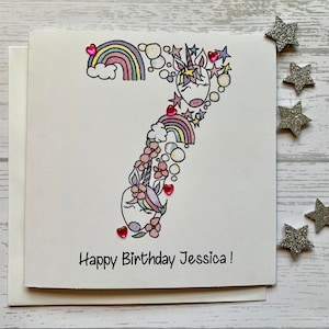Carte personnalisée du 7e anniversaire licorne. Illustration aquarelle de style dessin animé/griffonnage du chiffre 7 agrémentée de pierres précieuses en forme de coeur.