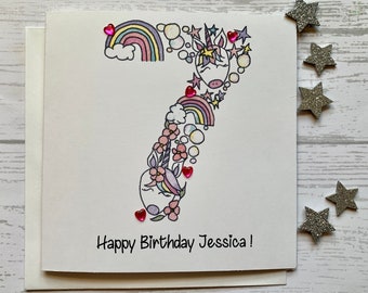 Carte personnalisée du 7e anniversaire licorne. Illustration aquarelle de style dessin animé/griffonnage du chiffre 7 agrémentée de pierres précieuses en forme de coeur.