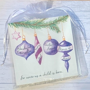 May include: A Christmas card with a purple, white, and green color scheme. The card features a watercolor illustration of ornaments with the words "Wonderful Counselor", "Mighty God", "Everlasting Father", and "Prince of Peace" written on them. The card also includes the text "For unto us a child is born..."