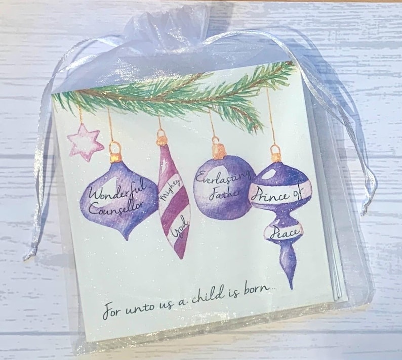 May include: A Christmas card with a purple, white, and green color scheme. The card features a watercolor illustration of ornaments with the words "Wonderful Counselor", "Mighty God", "Everlasting Father", and "Prince of Peace" written on them. The card also includes the text "For unto us a child is born..."