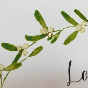 May include: A white card with a green mistletoe sprig and a gold glitter embellishment. The card says "Love" in black script.