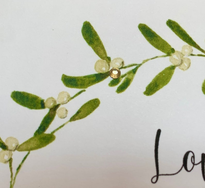 May include: A white card with a green mistletoe sprig and a gold glitter embellishment. The card says "Love" in black script.