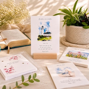 Peut inclure: Plusieurs cartes de motivation avec des illustrations à l'aquarelle. L'une représente un phare, une autre un champ de fleurs et une troisième un goéland sur une plage. Les cartes sont présentées avec un livre ouvert et une plante en pot.