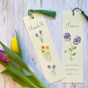 Può includere: Due segnalibri color crema con nappe verdi. Uno presenta il nome "Elisabeth" e illustrazioni floreali. L'altro ha "Claire" e una citazione da Matteo 6:28. Un mazzo di tulipani è visibile.