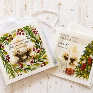 May include: Two square greeting cards featuring watercolour illustrations of a manger scene. The text reads, "OTHERS SEE CHRISTMAS... But we see JESUS! HEBREWS 2:9." One card shows a pine wreath with red berries, while the other shows a Christmas tree.