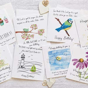 May include: A collection of eight square greeting cards featuring watercolor illustrations of birds, flowers, cupcakes, and a lighthouse. Each card includes handwritten text, scripture verses, and small wooden embellishments attached to the paper surface.