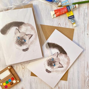 May include: Watercolor greeting cards featuring a Siamese cat with blue eyes. The cat is depicted looking upwards. The cards are packaged with brown envelopes. Paint tubes and a paintbrush are visible in the background.
