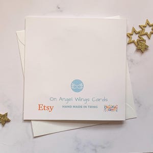 May include: A white card with the text "On Angel Wings Cards" and "Etsy" in orange. The card also has a blue logo with wings and the text "Hand Made in Tring" and "Cheerfully Given" in a colorful font.