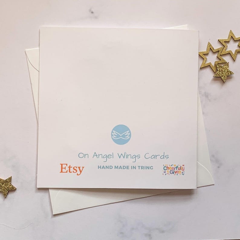 May include: A white card with the text "On Angel Wings Cards" and "Etsy" in orange. The card also has a blue logo with wings and the text "Hand Made in Tring" and "Cheerfully Given" in a colorful font.