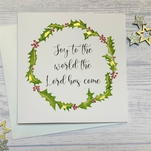 May include: A white card with a green holly wreath and red berries. The text inside the wreath reads "Joy to the world the Lord has come".