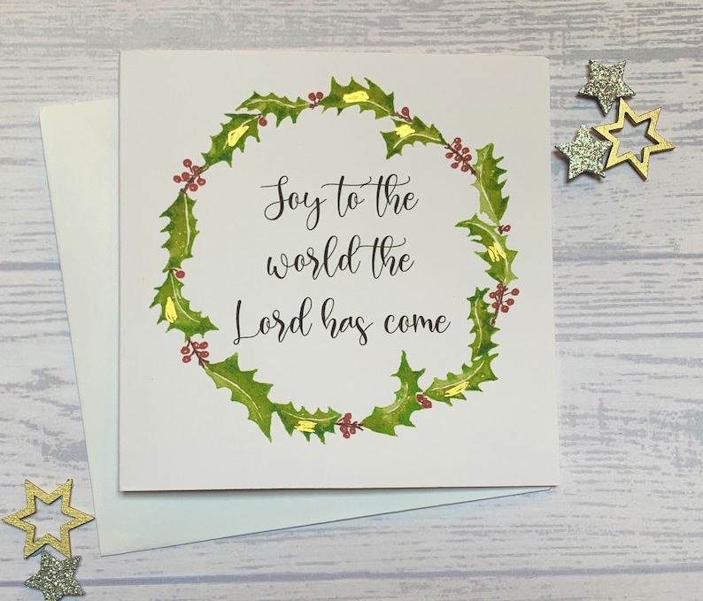 May include: A white card with a green holly wreath and red berries. The text inside the wreath reads "Joy to the world the Lord has come".