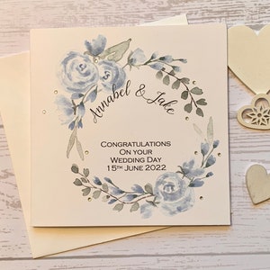 Carte de mariage personnalisée avec un cercle de fleurs bleu poudré et de délicats strass. Félicitations. Le jour de votre mariage