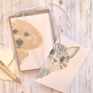 May include: A set of greeting cards with watercolour portraits of a dog and a cat. The dog is light brown, and the cat is grey with black stripes. The cards are packaged in a sheer white bag, with a wooden pen and heart-shaped decoration.