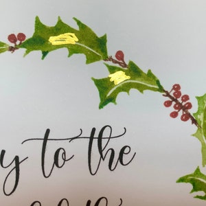 May include: A Christmas card with a holly wreath and gold accents. The text on the card reads "Joy to the".