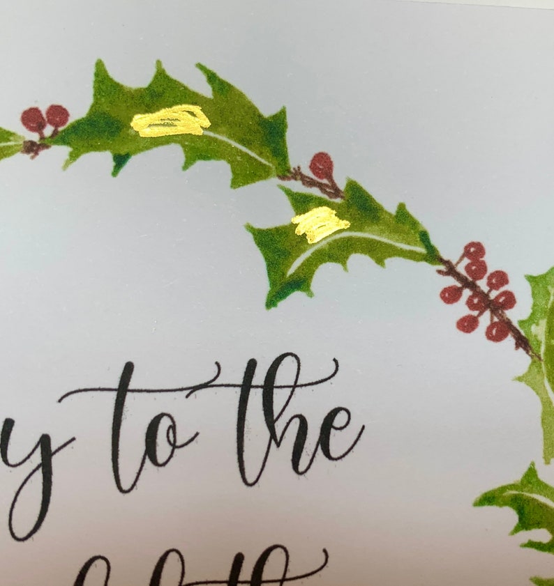 May include: A Christmas card with a holly wreath and gold accents. The text on the card reads "Joy to the".