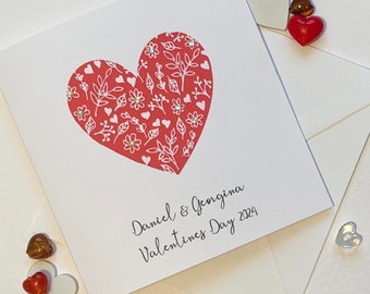 May include: A white Valentine's Day card featuring a red heart filled with white floral and leaf patterns. The card is inscribed with "Daniel & Georgina Valentines Day 2024". Decorative heart-shaped embellishments are placed around the card.