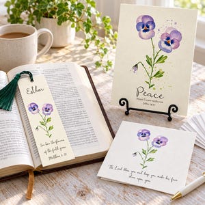May include: A bookmark with a dark green tassel, a vertical greeting card on a black stand, and a square folded card, all featuring watercolour pansy illustrations. The bookmark reads Esther and Matthew 6:28, the vertical card reads Peace John 14:27, and the square card features a blessing.