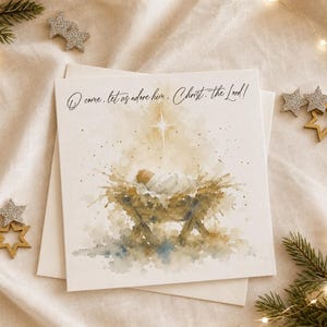 May include: A square greeting card featuring a watercolor illustration of a baby in a manger under a bright star. The text O come, let us adore him, Christ, the Lord! is printed in black script at the top against a white background.