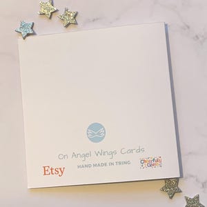 May include: A white card with blue wings and the text "On Angel Wings Cards" in black. Below the wings is the text "Etsy" in orange and "HAND MADE IN TRING" in black. In the bottom right corner is a colorful square with the text "Cheerfully Given".