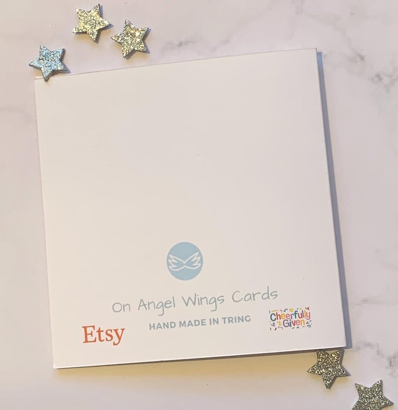 May include: A white card with blue wings and the text "On Angel Wings Cards" in black. Below the wings is the text "Etsy" in orange and "HAND MADE IN TRING" in black. In the bottom right corner is a colorful square with the text "Cheerfully Given".