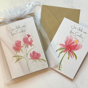 May include: Two floral greeting cards with watercolour peony illustrations. One card is inside a sheer white gift bag. The cards have the text "Just a little note from Caroline" in script. A brown envelope is behind the cards.