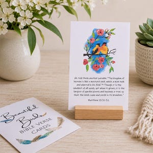 May include: A white rectangular card featuring a watercolour illustration of two yellow and black birds perched on a branch with red flowers and blue accents, with the text of Matthew 13:31-32 printed below, displayed on a small wooden block stand.