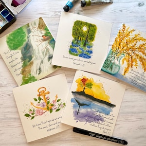 May include: A collection of square watercolour paintings depicting nature scenes, including trees and flowers. Each card features a Bible verse. A black Micron Graphic pen is also visible. The cards are on a wooden surface.