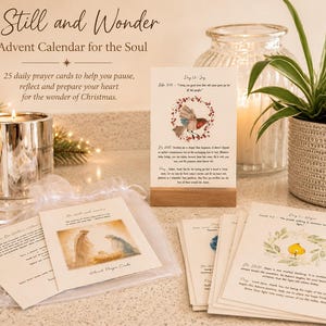 May include: A set of 25 daily prayer cards titled Be Still and Wonder, An Advent Calendar for the Soul. The cards feature watercolor illustrations of a robin and a candle, with printed text, displayed on a wooden stand and in a sheer bag.