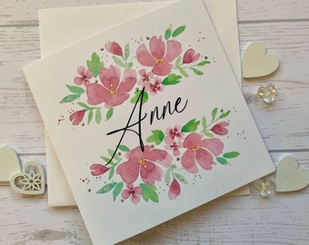 Carte d'anniversaire prénommée. Des fleurs roses lâches à l'aquarelle et des pierres précieuses délicates entourent le nom de votre destinataire.