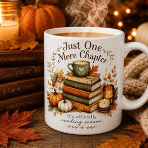 May include: A white ceramic mug featuring a graphic of a stack of books, pumpkins, and a small green cup. The text reads Just One More Chapter and It's officially reading season. The design includes autumn leaves and candles in warm colors.