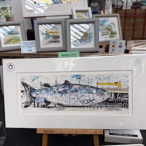 May include: A framed art print depicting a blue and white fish design with architectural details. The print is in a white frame, with other framed artworks visible. The print is labeled "Seaham & Newcastle (Fish)". The print is displayed on a wooden easel.
