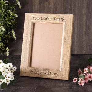 May include: A light-colored wooden picture frame with a visible grain pattern. The top border is engraved with the text "Your Custom Text" followed by a heart symbol, and the bottom border features the text "Engraved Here" with a heart.