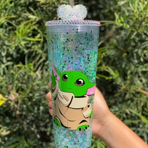 May include: A clear tumbler with a green and brown cartoon character on it. The tumbler is filled with iridescent glitter and has a white heart-shaped lid with rhinestones.
