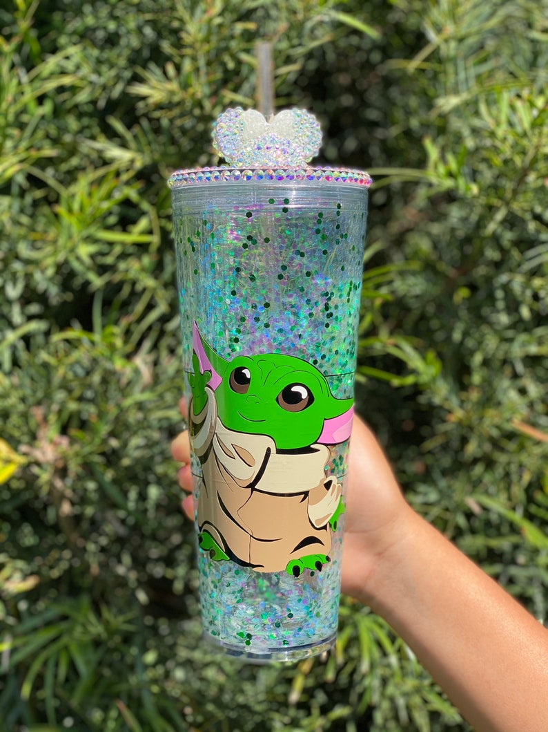 May include: A clear tumbler with a green and brown cartoon character on it. The tumbler is filled with iridescent glitter and has a white heart-shaped lid with rhinestones.