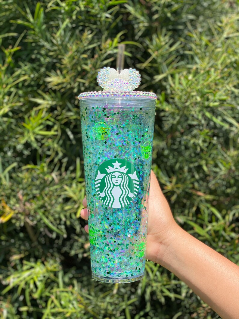 May include: A clear plastic tumbler with a green glitter design and a white heart-shaped lid with rhinestones. The tumbler features a green Starbucks logo and a green Yoda character.
