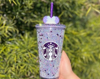May include: A clear reusable cup with a purple and white heart lid and a purple straw. The cup is covered in iridescent glitter and has a purple Starbucks logo with a crown on top.