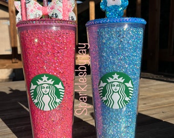 May include: Two glitter-filled tumblers, one pink and one blue, with Starbucks logos. The pink tumbler has a bow-shaped lid, and the blue one has a Mickey Mouse-shaped lid. Both have straws.