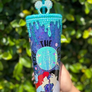 May include: A clear plastic tumbler with a purple and blue glitter base. The tumbler has a blue and white glitter rim and a blue and white glitter drip design. The tumbler features a black and white Starbucks logo with the text "The Kiss" and a cartoon image of Ariel and Prince Eric from The Little Mermaid.