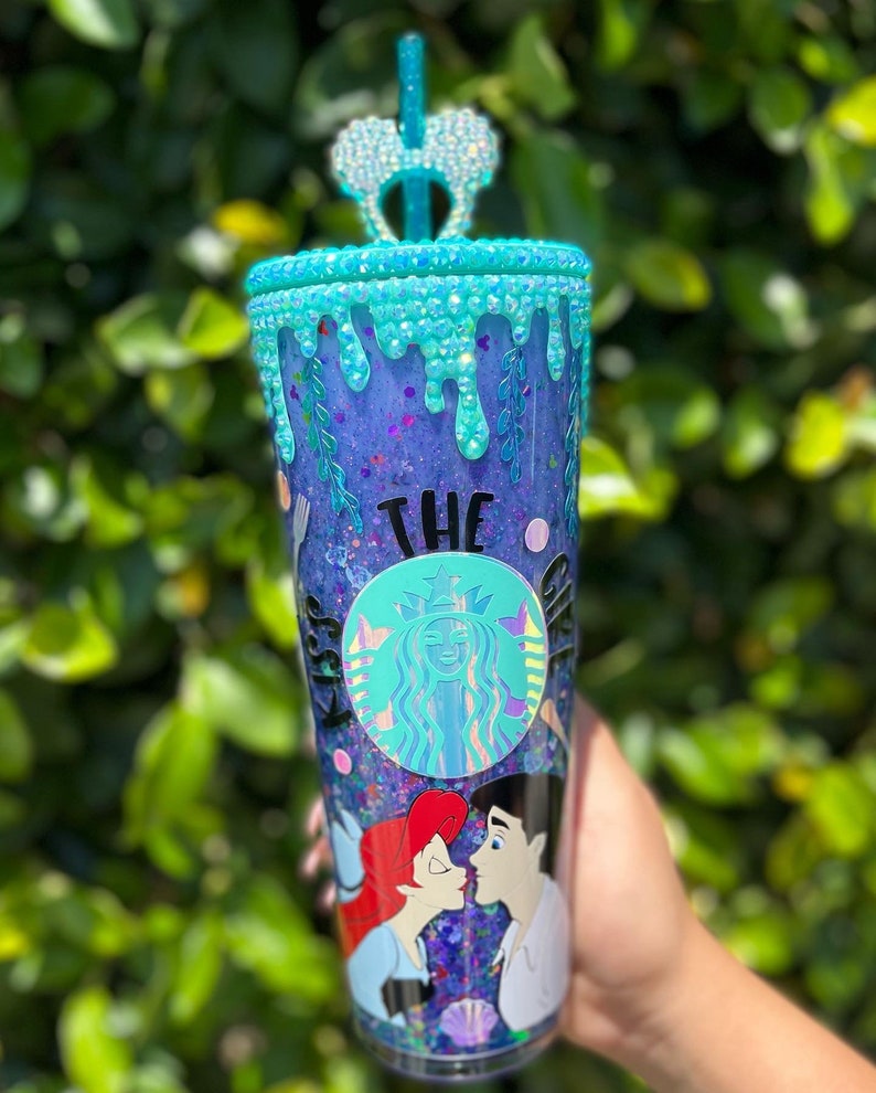 May include: A clear plastic tumbler with a purple and blue glitter base. The tumbler has a blue and white glitter rim and a blue and white glitter drip design. The tumbler features a black and white Starbucks logo with the text "The Kiss" and a cartoon image of Ariel and Prince Eric from The Little Mermaid.