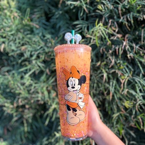 Puede incluir: Un vaso de glitter naranja con un dise&ntilde;o de Minnie Mouse. Minnie lleva un vestido marr&oacute;n y sostiene una taza de caf&eacute;. El vaso tiene una tapa transparente y una pajita plateada.