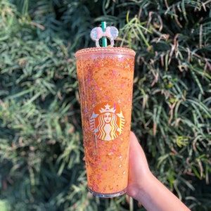 Puede incluir: Un vaso reutilizable transparente con un dise&ntilde;o brillante naranja y dorado. El vaso tiene el logotipo de Starbucks y un adorno de pajita de orejas de Minnie Mouse rosa.