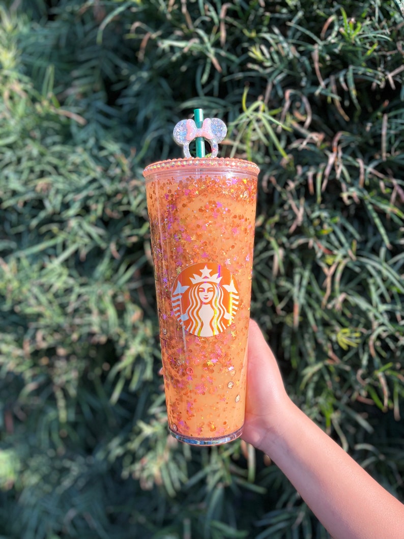 Puede incluir: Un vaso reutilizable transparente con un dise&ntilde;o brillante naranja y dorado. El vaso tiene el logotipo de Starbucks y un adorno de pajita de orejas de Minnie Mouse rosa.