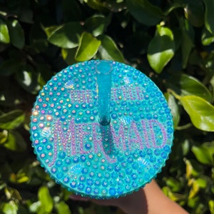 May include: A turquoise blue reusable cup with a clear lid and straw. The lid is covered in iridescent rhinestones and the word "Mermaid" is spelled out in rhinestones.