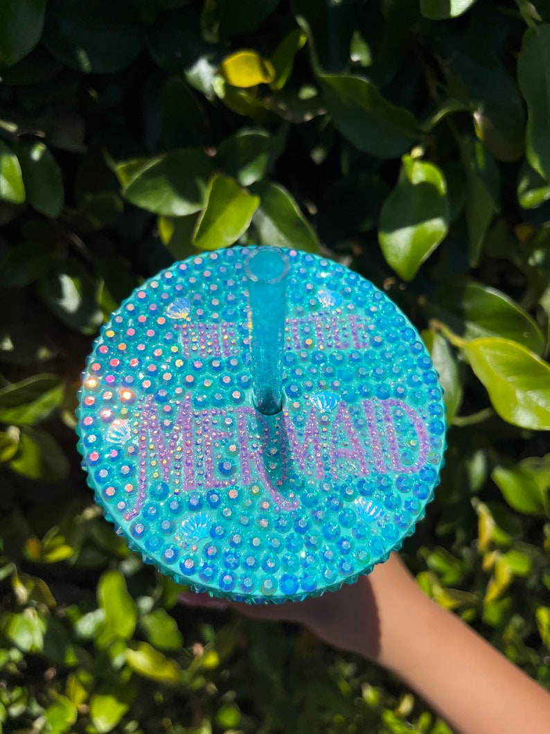 May include: A turquoise blue reusable cup with a clear lid and straw. The lid is covered in iridescent rhinestones and the word "Mermaid" is spelled out in rhinestones.