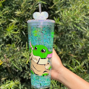 May include: A clear tumbler with a green glitter design and a character from the Star Wars franchise, Baby Yoda, printed on the side. The tumbler has a clear lid with a heart-shaped rhinestone embellishment and a clear straw.