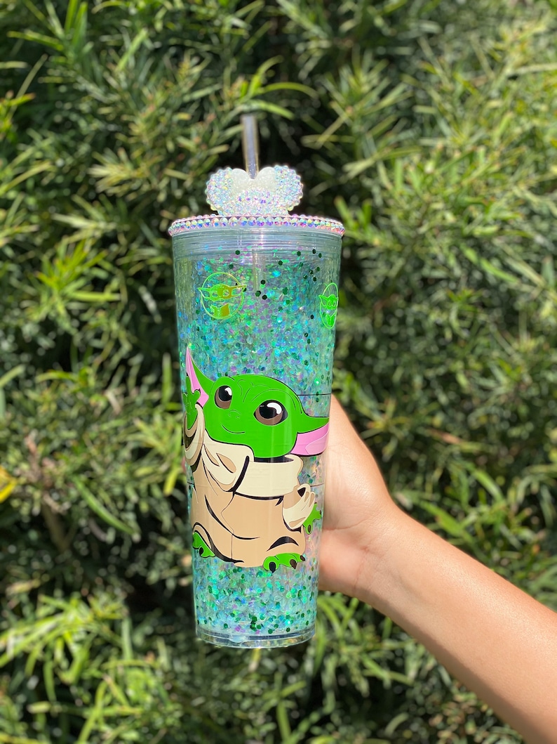 May include: A clear tumbler with a green glitter design and a character from the Star Wars franchise, Baby Yoda, printed on the side. The tumbler has a clear lid with a heart-shaped rhinestone embellishment and a clear straw.