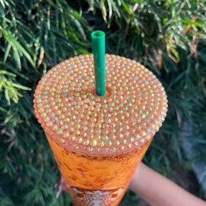 Puede incluir: Un vaso alto y transparente con un dise&ntilde;o brillante naranja y dorado. El vaso tiene una tapa con una pajita verde y est&aacute; cubierto de pedrer&iacute;a iridiscente. El logotipo de Starbucks es visible en el lateral del vaso.