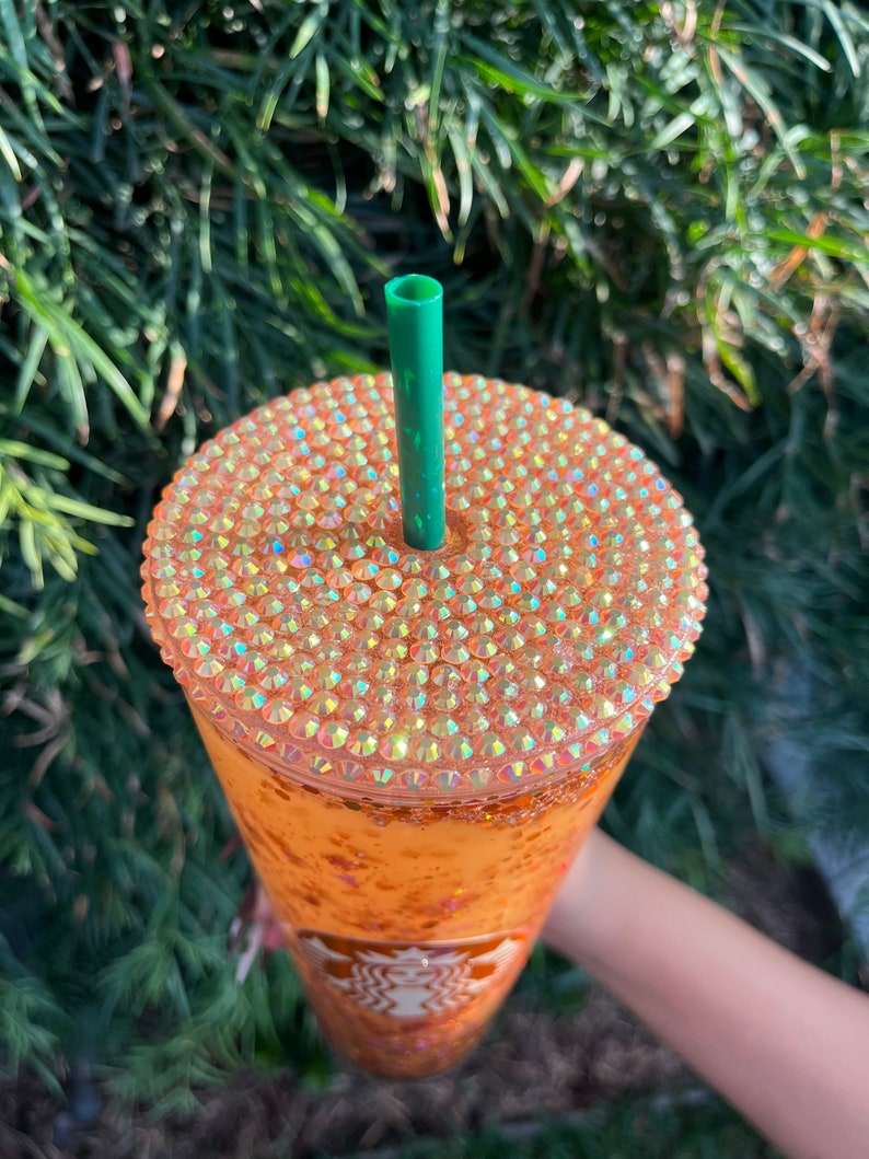 Puede incluir: Un vaso alto y transparente con un dise&ntilde;o brillante naranja y dorado. El vaso tiene una tapa con una pajita verde y est&aacute; cubierto de pedrer&iacute;a iridiscente. El logotipo de Starbucks es visible en el lateral del vaso.