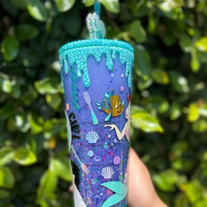 May include: A purple and blue glitter tumbler with a mermaid theme. The tumbler has a blue and green dripping design, a fork and knife, a seashell, and a fish. The tumbler also has a mermaid tail and the text "Ariel" on the side.